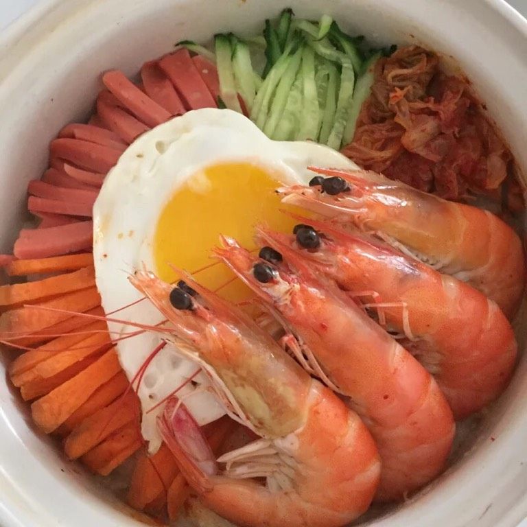 石锅拌饭,如图砂锅空锅上火加热,四周抹油,倒入米饭,用铲子轻轻压平,放入黄瓜,胡萝卜,火腿,辣白菜,鸡蛋和虾