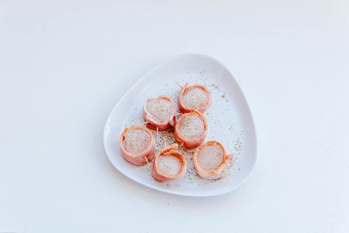 培根扇贝卷,将扇贝卷在<a style='color:red;display:inline-block;' href='/shicai/ 443'>培根</a>内，用牙签稍作固定，<a style='color:red;display:inline-block;' href='/shicai/ 443'>培根</a>长短宽窄根据扇贝的大小需要做一下切边处理，定型后双面撒黑<a style='color:red;display:inline-block;' href='/shicai/ 724'>胡椒</a>备用