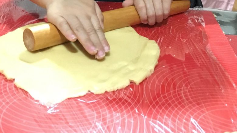 如何做出酥脆的卡通饼干,将面团放在案板上摁扁,在上面铺上一层保鲜膜,用擀面杖擀成0.3cm厚度的面饼,打开保鲜膜,开始用模具制作
