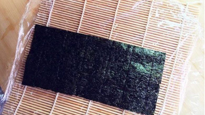 樱花寿司,首先将寿司帘上铺上保鲜膜,再放上一片海苔。