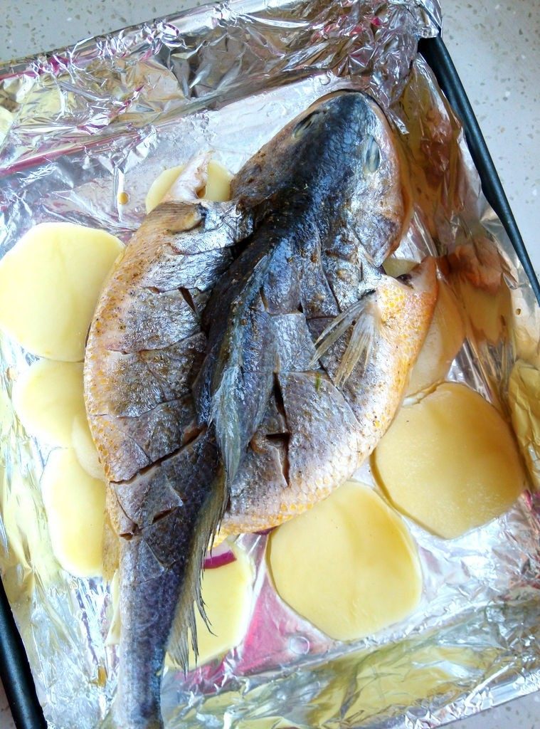 香辣豆豉烤全鱼,烤盘内铺锡纸,锡纸上刷油,将切好的土豆片铺在底层。把腌好的鱼放在上面,然后倒入刚才的腌料