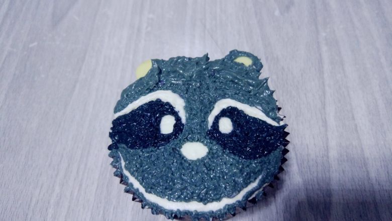 小浣熊立体彩绘杯蛋糕,将白巧克力片插在杯蛋糕内,充当耳朵,在上面外圈挤上浅灰色,内圈是深灰色奶油霜