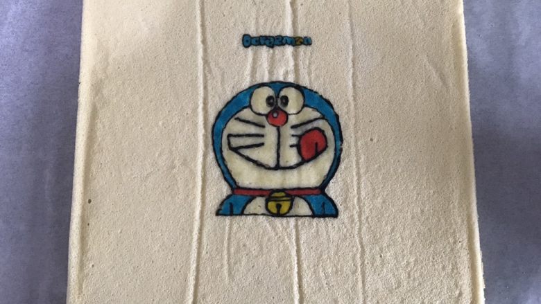 哆啦A梦彩绘蛋糕卷,放至蛋糕卷表面不再冒热气,用手将蛋糕卷四周与模具剥离,表面铺上油纸,倒扣烤盘脱模。
轻轻将油纸撕去,轻轻哦,不要破坏图案。
看,蓝胖纸在对你笑哦😜
铺上油纸,放凉。