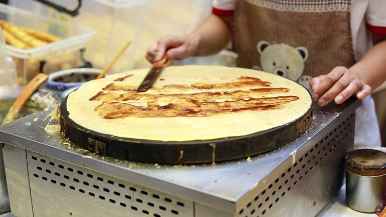 天津老味煎饼果子,这时小铲子登场,用小铲子轻轻松动煎饼边缘,双手一提一翻,煎饼就被反过来了,顿时能闻到煎饼的清香味,蛋香味,香葱味,在煎饼上抹上少许的天津甜面酱,腐乳酱汁和辣椒酱,煎饼上一道深褐色,一道浅粉色,一道酱红色,咸香甜俱全。
