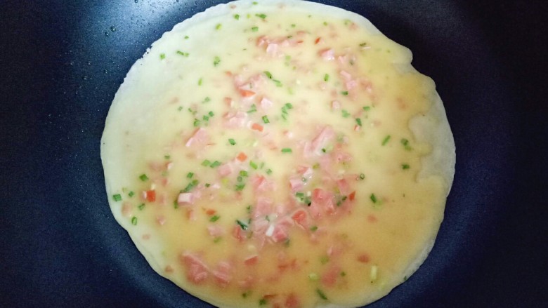 火腿鸡蛋饼,热锅中,盛两勺面糊,拿起平底锅摇晃一圈,再用勺子底部将面糊摊平。
小火煎至鸡蛋饼表面的蛋液凝固即可。