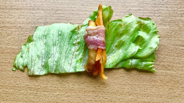 薯条的N种吃法,当培根卷起边就可以出锅了。然后如图先横放着生菜,再上面横放培根,放薯条和叮好的胡萝卜丝,卷起培根。