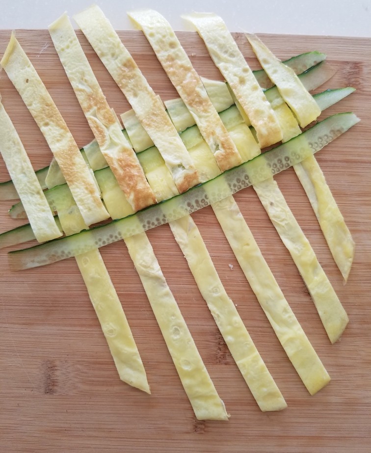 菠萝蛋包饭,把鸡蛋条和黄瓜条按照编花篮的方法编成蛋饼,如图每隔一个把鸡蛋条揭起放入黄瓜条。看到揭开蛋条贴着锅底的颜色了吧,这就是为什么煎蛋饼时不要翻面,要保证表面颜色漂亮,翻面贴到锅底就会有斑纹。