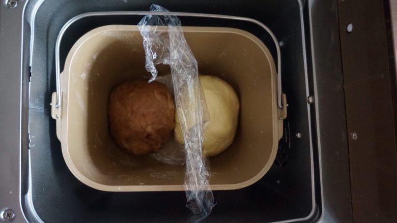 面包机版双味吐司,将面包桶里的搅拌刀取出,将两份面团滚圆后放入,中间放入保鲜膜隔开,选择面包机的发酵程序40分钟