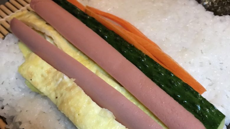 寿司卷,将适量米饭放在事先铺好的寿司帘的海苔上面,带上一次性手套按压结实,最上面留出一些空间。