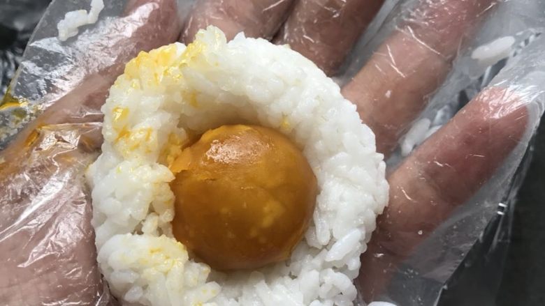 咸蛋黄肉松饭团-泰迪版,手戴一次性手套取一小把温热的饭团(能包褒蛋黄不漏,或把蛋黄压碎和饭团和一起也行),稍捏紧按扁,放入一颗蛋黄;