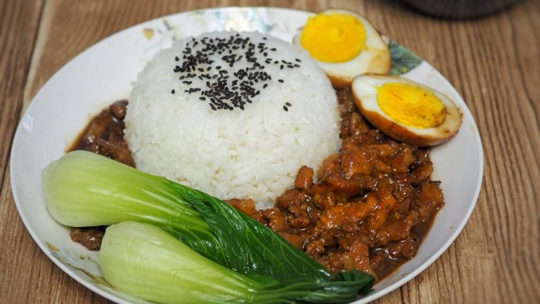 台湾卤肉饭,取一小碗,碗底撒少量熟芝麻,盛饭后倒扣在盘子中,周围淋上卤肉,铺上青菜,摆上鸡蛋。