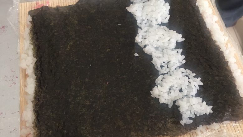 翻转樱花寿司,在把紫菜翻过来,撒有樱花粉米饭朝下,光滑紫菜面朝上,在靠自己方向三分之一处铺一些米饭