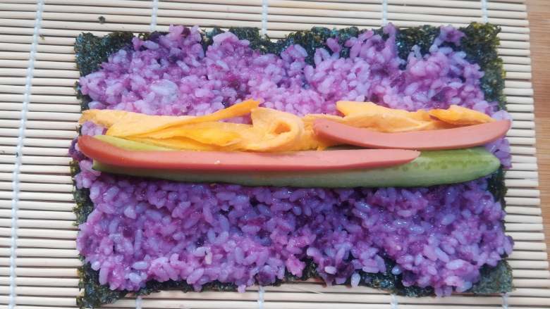 紫薯寿司,把拌匀的紫薯米饭均匀的铺在紫菜上,紫菜的两端也要有米饭粒。如果觉得米饭比较粘,可以用勺子沾水,有勺背来把米饭推均匀。用手来操作的话,也可以在手上抹些凉开水,或寿司醋防止黏手。