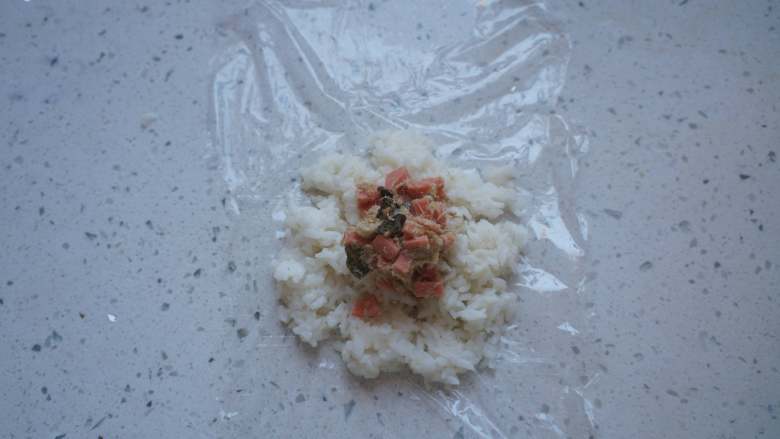 小猪饭团,保鲜膜刷一层食用油,铺上适量米饭,放一勺香肠肉松,借助保鲜膜做成饭团。