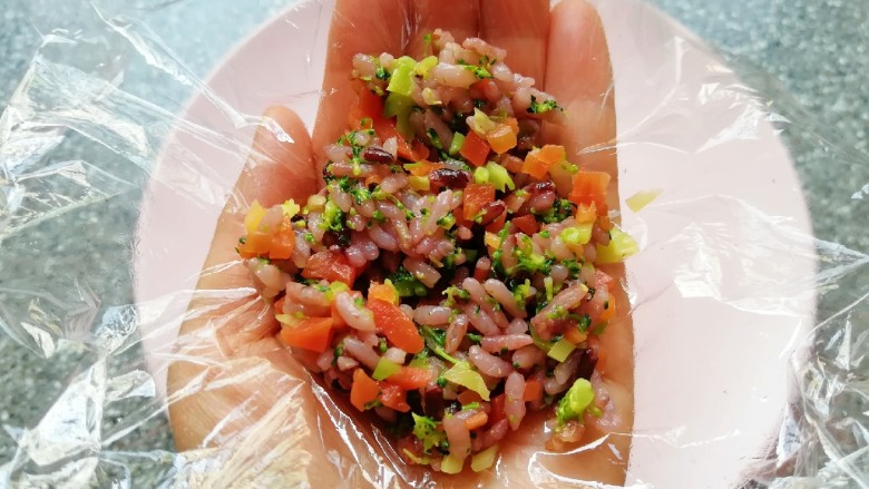 杂粮蔬菜饭团,手掌心里放上一张保鲜膜,舀入拌好的杂粮蔬菜饭,提起保鲜膜的四个角,用手捏好饭团。