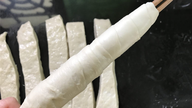 烤面筋,准备好两根筷子,取一条面筋,切面朝上,一头用筷子夹住后用手慢慢抻开面团,一手转动筷子让面团缠在筷子上,一层错开压住一层,收口处捏紧,防止脱落。