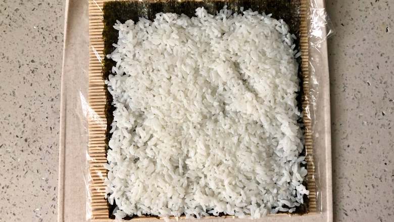 萌萌哒“小蜜蜂寿司”,拯救宝宝的味蕾,寿司席上可以先包一层保鲜膜,这样方便清洗。把海苔铺在寿司席上,先铺上一层米饭。米饭拌好寿司醋后要凉透后再铺到海苔上,否则太湿热海苔会起皱,而且不好切。