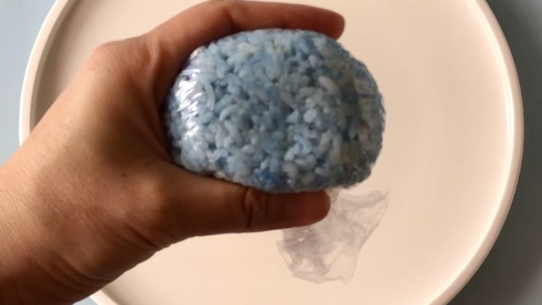 六一儿童节到啦,给孩子做一份萌萌的小河马卡通早餐吧!,取一张保鲜膜,把蓝色米饭放在保鲜膜中间,然后借助保鲜膜把米饭裹起来,捏出河马的头。