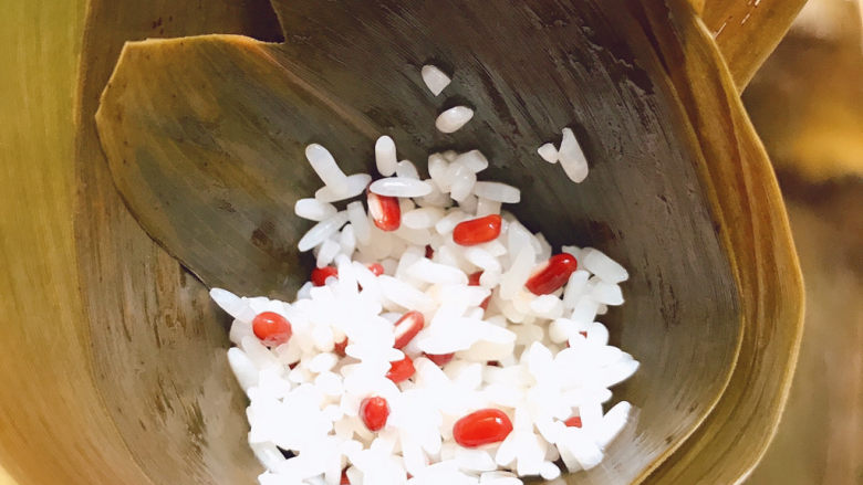 红豆粽子,拿两片<a style='color:red;display:inline-block;' href='/shicai/ 211314'>粽叶</a>,毛面向下卷成锥形,放入一勺糯米