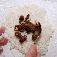 蜜汁小卷三角饭团,撕开一层保鲜膜为底,将<a style='color:red;display:inline-block;' href='/shicai/ 5188'>寿司饭</a>平均铺成圆形,取适量炒小卷放在饭中间。
由于炒过的小卷热气遇饭容易出水,建议可将炒小卷放凉后再包。