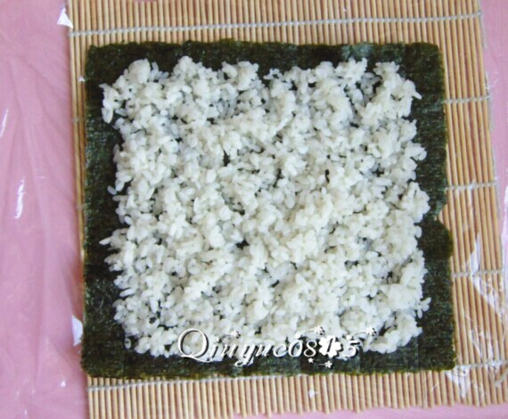 翻转樱花寿司,寿司帘上铺一张保鲜膜,放海苔,把米饭铺匀。海苔其中一端留出一点空边