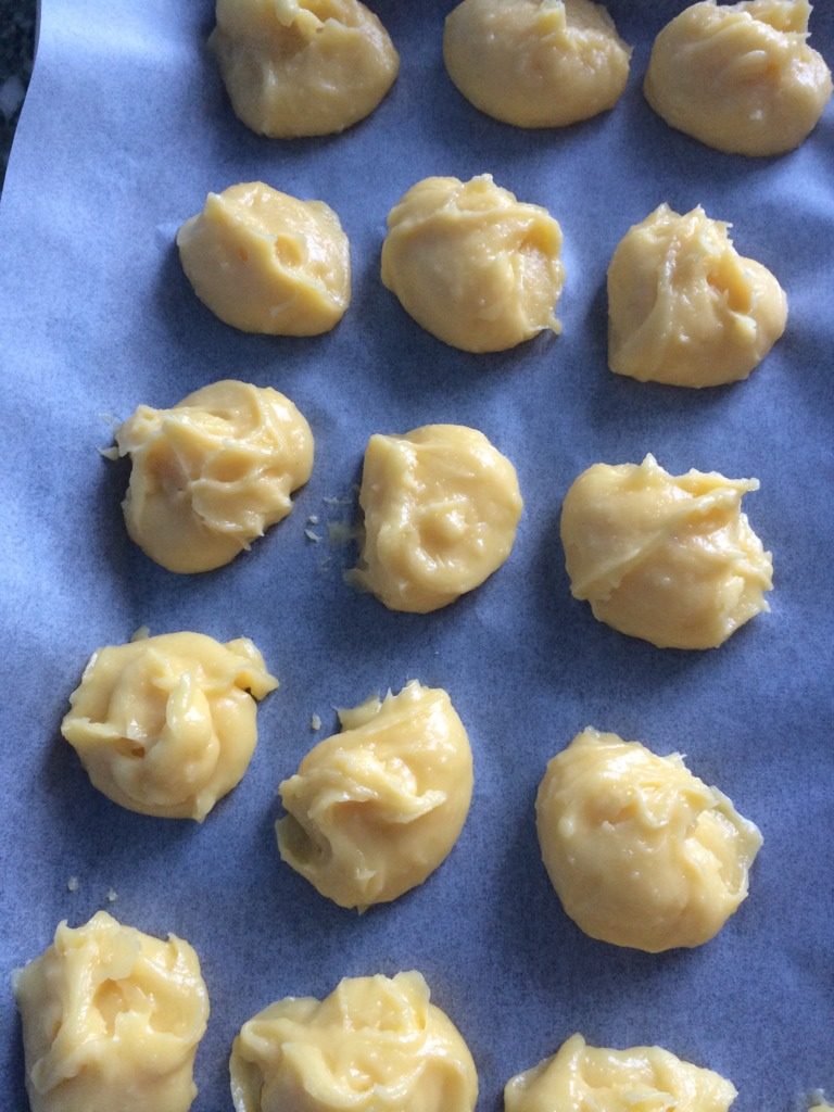 泡芙球球,用匙舀起一块混合物,倒在铺好油纸的烤盘上。用手指蘸水,压去顶部的小尖,使其圆圆的。